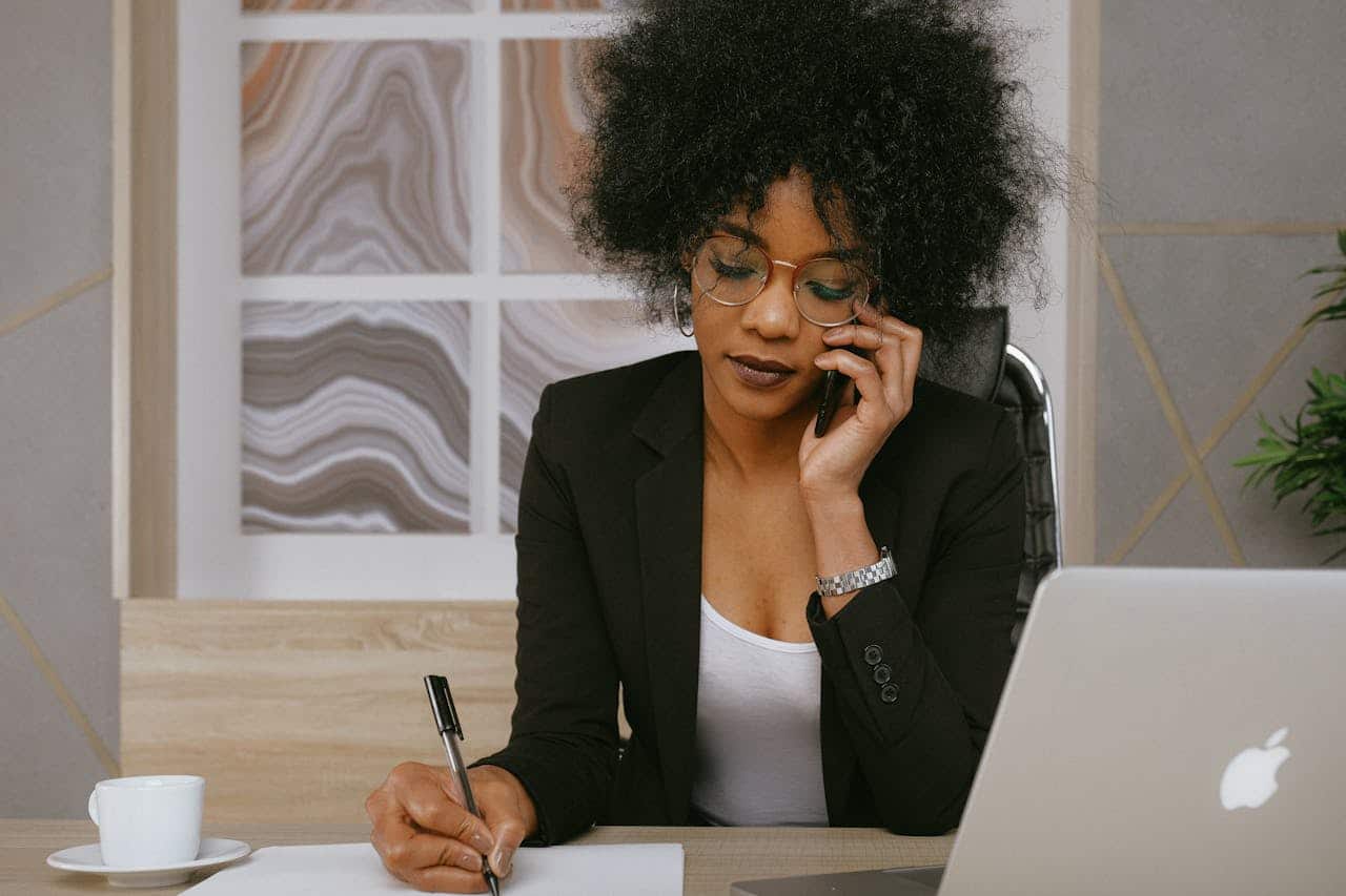 Donna al telefono in ufficio mentre prende appunti, operatrice call center con laptop e tazza di caffè.